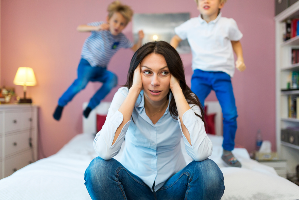 Erschöpfte Mutter sitzt auf dem Bett, während Kinder im Hintergrund spielen
